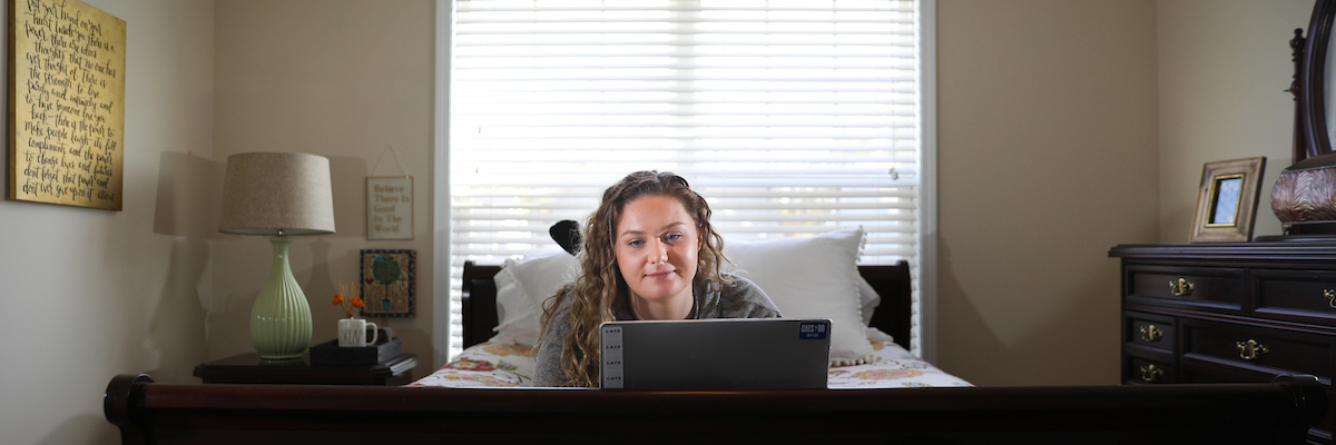 A white female student works on a laptop while laying in bed.