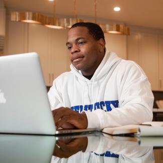 A black male student wearing a UK sweatshirt sits at a dining room table working on a MacBook.